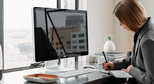 A woman taking notes at a desk in front of a computer monitor